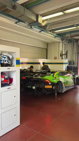 The video shows two individuals wearing racing suits working on a green and yellow Lamborghini race car in a garage. The car is elevated on stands, with its rear wheels removed. The hood of the car is open, exposing the engine. The garage appears to be well-lit with fluorescent lights. To the left, a white cabinet displays two racing helmets. The floor is red. One person is near the rear of the car, while the other is on the right side, possibly interacting with equipment or the car. The overall scene suggests a pit stop or maintenance operation for a racing vehicle.