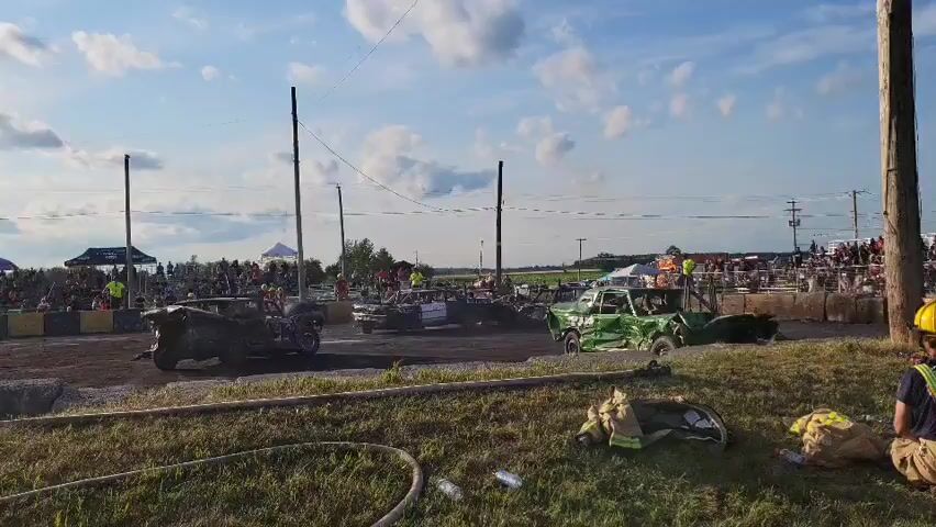The video shows a demolition derby event on a sunny day with a blue sky and scattered clouds. In the foreground, a person wearing a yellow helmet and a reflective vest kneels on grass, with firefighting equipment nearby. The main action is in the background on a dirt track, where several heavily damaged cars are present. One green car is particularly mangled. Spectators are visible behind a barrier. Two tall poles with wires strung between them stand on the track. The overall scene depicts an active demolition derby event.