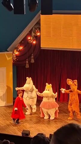 A stage performance features three costumed characters and one person in a red outfit. Two of the costumed characters are white, hippopotamus-like figures, one wearing a suit and the other a pink tutu. The third costumed character is a brown, rabbit-like figure. A person in a bright red dress with a black belt and black shoes is dancing on stage, performing a high kick. The stage has a wooden floor and a red curtain backdrop. To the left, there is a yellow wall with a white door. A large orange screen is visible above the stage. Several audience members are visible in the foreground, and stage lighting fixtures are mounted overhead.