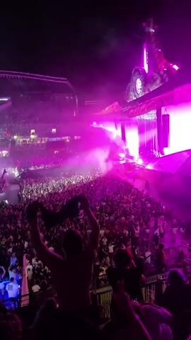 A night time aerial shot of a large outdoor concert. The crowd is packed into a stadium area, with a stage at one end. The stage is elaborate, with bright lights and pyrotechnics. A haze or smoke hangs over the crowd, illuminated by the stage lights. People in the crowd are cheering and raising their hands. The stadium seating is visible in the background, also filled with people.