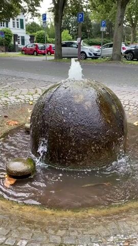 This is a close-up, medium shot video of a small, round fountain. The fountain is made of dark, textured material and has water spraying from the top. Water also trickles down the sides of the fountain and pools in the basin below. Two smaller, round stones sit in the water to the left of the main fountain element. The fountain is situated on a cobblestone surface. In the background, there is a street with cars parked along the side, some trees, and buildings. The sky is overcast.