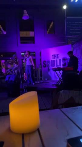 A live music performance is taking place on an outdoor stage at night. The stage is lit with purple and green lights. A female singer stands center stage, wearing a white tank top and dark pants, holding a microphone and singing. To her left, a male musician plays a trumpet. To her right, a male musician plays a keyboard. Behind them, a banner reads "THE SOULIST." In the foreground, a lit candle sits on a wooden table, with a blurry audience visible in the background.