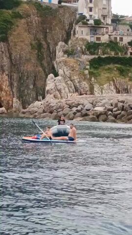 The video shows a serene coastal scene with a rocky cliff face and buildings perched on top. In the foreground, a man and a woman are on a paddleboard in the water. The man is sitting on the paddleboard, while the woman is lying down. The water is calm and a few ripples are visible. The cliff face is rugged and appears to have some vegetation growing on it. A few small buildings, likely houses or apartments, are visible on the top of the cliff. The scene suggests a peaceful day at the beach.