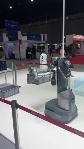 The video shows a robotic arm mounted on a mobile base, displayed at an exhibition. The robot is gray and silver and has a screen on its front. It is positioned next to a white shelving unit with gray crates on it. In the background, there are exhibition booths, people walking around, and signage. The floor is white with red rope barriers. There is another white robot in the background.