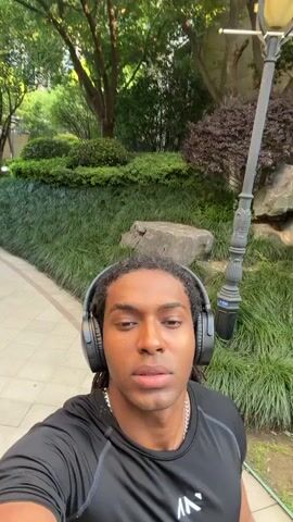 The video shows a person walking down a paved path in a garden. The person appears to be a man with dreadlocks, wearing headphones, a black athletic shirt with a white logo, and a silver chain necklace. He is holding the camera, recording himself as he walks. The garden has trees, bushes, and a lamp post. In the background, a tall building can be seen. The man smiles at the camera as he moves along the path.