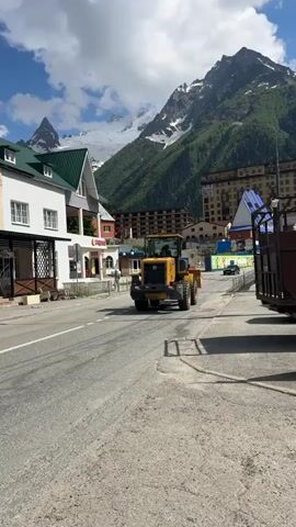 This media file captures a scenic view of a street in Dombay, Russia, with a prominent mountainous backdrop. 

**Subjects:** The primary subjects include a yellow loader vehicle, a black car, a few people walking on the sidewalk, and various buildings lining the street. The imposing mountains, partially covered in snow and lush green vegetation, dominate the background.

**Activity/Event:** The loader is seen moving down the street, suggesting ongoing construction or maintenance work. A car is also in motion, indicating normal daytime traffic. People are walking, suggesting a relaxed atmosphere in the town.

**Notable Details:** The time of day appears to be daytime, with bright sunlight casting shadows. The weather is clear with scattered clouds, and the presence of snow on the mountain peaks suggests a cooler climate or high altitude. The architecture of the buildings, with their pitched roofs and balconies, provides a clue to the local style. The overall emotion evoked is one of a peaceful, active mountain town.

**Visible Text:** "COCA-COLA" is visible on a sign above one of the buildings. Other text is too small or obscured to be legible.