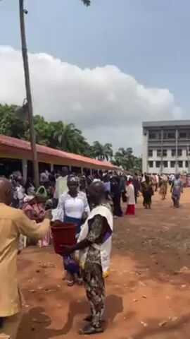 The video shows a large crowd of people, mostly wearing colorful clothing. Many people are moving around. There is a tall building in the background and some trees. A small structure is visible with people sitting on it. Some people are holding water bottles.