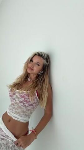 A young woman with blonde hair and a white lace top and white skirt leans against a white wall. She smiles and poses for the camera, touching her hair and hand at various points in the short video.