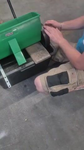 The video shows a person kneeling beside a green hopper with wheels. The hopper is positioned on top of a black container. The person's hands are visible, and they appear to be touching the hopper's wheel or some part of the mechanism. The person is wearing shorts with pockets. The environment appears to be indoors, possibly a garage or workshop, with a concrete floor. There are some wooden planks visible under the hopper. The hopper has a label that reads "GO ON." The person's actions suggest they are interacting with or adjusting the hopper, possibly preparing it for use or testing it.
