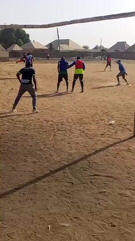 The video shows a soccer game being played on a dirt field. The players are wearing different colored shirts, including blue and red. They are running and kicking the ball. There are goals at each end of the field, and houses are visible in the background. The sky is bright.