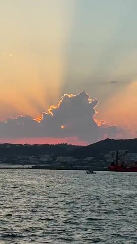 The video shows a sunset over the ocean. The sun's rays are visible through a cloud. There is a town and a hill in the background. There is also a boat in the water and a large orange structure that appears to be a crane or some kind of industrial equipment. The water is choppy.