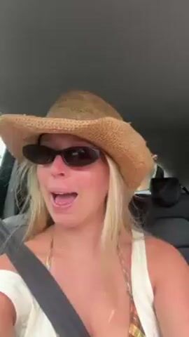 A woman with long blonde hair, wearing a straw cowboy hat, black sunglasses, and a white top over a bikini top, is sitting in the driver's seat of a car. She is wearing a seatbelt. She is looking at the camera and speaking with a wide open mouth, expressing excitement. The interior of the car is visible, including the rearview mirror and passenger seat. The car appears to be moving.