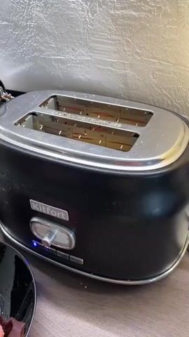 The video shows a black Kitfort toaster with two slices of bread inside. The toaster has a chrome top and a control dial with a blue light. The bread is being toasted, and the slices appear to be getting browner over time. The background is a textured white surface, possibly a wall or backsplash. A dark plate with some reddish food is partially visible in the lower left corner of the frame. The video appears to be a close-up shot of the toaster in action.