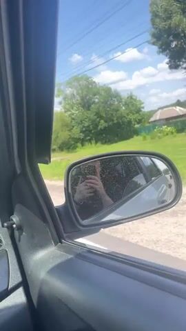The video shows a view from inside a car, looking out the side window. The car is on a dirt road, with a grassy area and trees on either side. In the distance, there are houses and utility poles. There are two traffic signs visible: one indicates no overtaking, and the other indicates a speed limit of 50. The sky is blue with some clouds. The reflection in the side mirror shows a person holding a phone, presumably recording the video. The overall scene suggests a rural or suburban setting on a sunny day.