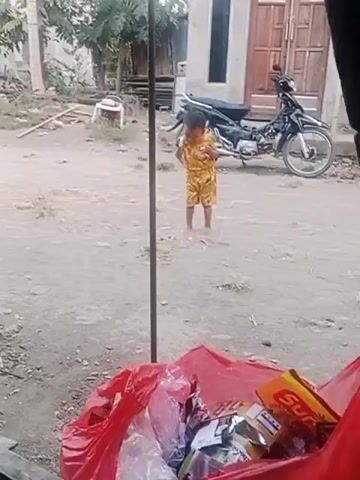 The video shows a young child standing in an outdoor area. The child is wearing a yellow outfit with a pattern. The child is facing the camera and appears to be holding something in their hands near their mouth. In the background, there is a motorcycle parked in front of a building. There is also a large red bag filled with trash in the foreground. The ground is dusty and uneven, with some sparse vegetation. The overall environment appears to be a rural or semi-rural setting.