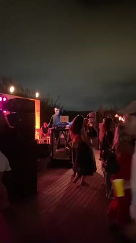 The video shows a group of people dancing at an outdoor event at night. The scene is illuminated by purple and pink lights. A DJ is visible behind a set of speakers on the left side of the frame. People are interacting and dancing on a wooden deck. The sky is dark and appears cloudy.