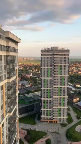 A vertical drone shot begins centered on a tall, gray apartment building with green accents. The drone then pans right, revealing a neighboring apartment building with yellow accents. In the background, a sprawling suburban landscape unfolds, dotted with houses and trees. The sky is a soft mix of blue and orange, with scattered clouds, suggesting the late afternoon or early evening. The camera continues to pan right, showing more of the city and the sky.
