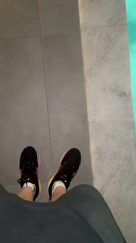 The video opens with a point-of-view shot looking down at two feet wearing black athletic shoes with red and yellow accents, walking on grey tiled pavement. A section of turquoise pool water is visible to the right. The camera then moves to the pool edge and enters the water. Inside the illuminated turquoise swimming pool at night, two individuals are visible. One person, with short dark hair, is seen from the chest up, looking upward and appearing to gasp or speak. A second person is partially submerged further back, with their hands near their face. The camera then pans left across the pool, showing the dark outdoor pool area with trees and distant lights from surrounding structures, before moving past the pool edge to reveal a pink bag and a skateboard resting on the dark tiled ground.