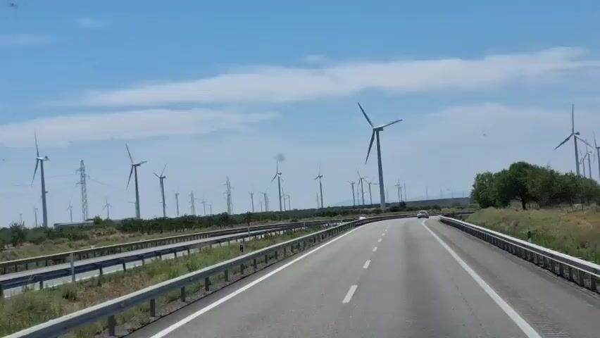 A shot from a vehicle traveling on a highway. The highway is divided by a median with guardrails. The landscape on either side of the highway is flat and dry with some sparse vegetation and numerous wind turbines scattered across the scene. The sky is blue with scattered clouds. Cars can be seen on the highway.