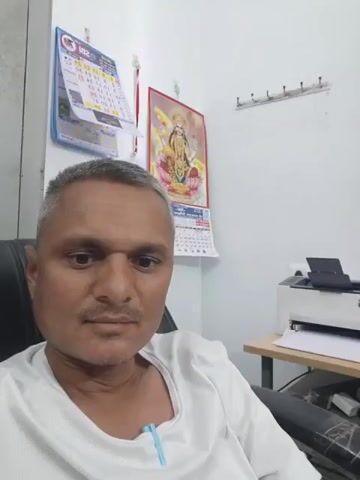 A middle-aged man sits in a chair, looking directly at the camera. He is wearing a white shirt. Behind him is a wall with two calendars and a row of coat hooks. To his right is a desk with a printer, a computer monitor, a keyboard, a mouse, and a speaker. The computer monitor displays a dark scene. The camera slowly pans to the right, focusing on the desk and the computer monitor before panning back to the man. A patterned curtain hangs to the right of the desk.