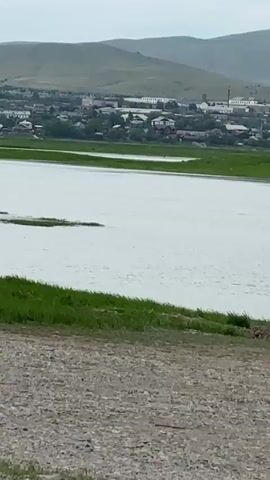 A video shows a body of water in the foreground with a green grassy area to the left. In the background, there is a town with buildings and houses, and rolling hills beyond. The sky is overcast. The video is shaky, suggesting it was recorded handheld. There are no people or animals visible in the video. The time stamp on the video indicates it was recorded on 00:00:00 to 00:00:04.