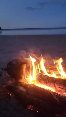 A close-up shot shows a campfire on a sandy beach at dusk. Logs are burning brightly, with flames and smoke rising. A metal mug sits to the left of the fire, appearing to be heated by the flames. In the background, a calm body of water stretches towards the horizon under a dark blue sky with a few clouds. The overall scene evokes a sense of peaceful solitude.
