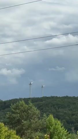 This media file depicts a landscape scene with two wind turbines visible on a distant, tree-covered hill. The sky is overcast with dramatic, dark clouds, suggesting stormy weather or the approach of a storm. In the foreground, lush green trees and foliage dominate the view, with some thinner, taller trees reaching upwards. Electrical wires are visible in the upper portion of the frame, spanning across the scene.

The camera appears to be moving, as indicated by the slight shift in perspective between the frames. In the foreground, a road sign with multiple white arrows pointing upwards on a blue background is partially visible on the right side of the frame. The overall impression is one of a rural or semi-rural setting, possibly in Ukraine, given the location context. The weather contributes to a somewhat moody and dramatic atmosphere, contrasting with the steady presence of the wind turbines. There are no people visible in the media. The time of day is not explicitly clear due to the cloudy conditions, but it appears to be daytime.