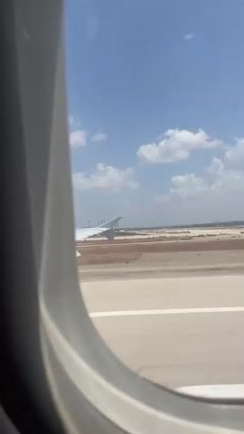 The video begins with a view out an airplane window. An airplane wing is visible in the foreground, and a runway stretches out in the distance. The sky is blue with scattered clouds. The camera then pans to the interior of the airplane, showing a seatback and an in-flight safety information card. The card is upside down and appears to be from a Turkish Airlines A320. The video ends with a close-up of the safety card.