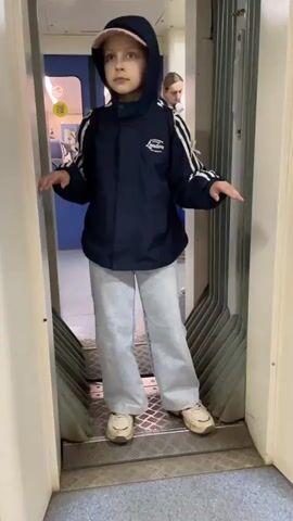A young girl in a navy blue jacket with a hood, light wash jeans, and tan sneakers is standing in the doorway between two train cars. She has her arms extended to the sides. The doorway is enclosed with white walls, and a series of horizontal silver metal strips line the floor. Behind her, through a blue door, other passengers can be seen in the train car. The train appears to be in motion.