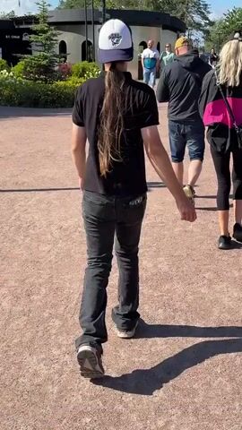 The video shows a person with long hair wearing a black shirt, jeans, and a white baseball cap walking away from the camera. They are on a gravel path with other people in the background. The environment appears to be outdoors, possibly a park or a public area, with some buildings and greenery visible. The lighting suggests it is daytime, with shadows cast on the ground. The video captures the person from behind as they walk, with the focus on their movement and appearance.