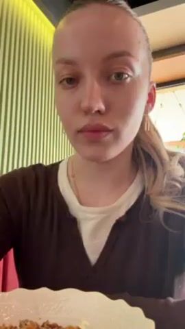 The video shows a young woman eating. She has fair skin, blonde hair pulled back in a low ponytail, and is wearing a brown sweater over a white shirt. She is sitting at a table in a restaurant, with green and yellow striped wall panels visible in the background. She eats something off of a white plate with a fork, looking into the camera. She chews with her mouth closed, and then looks off to the side.