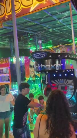 The video shows a group of young people at what appears to be a carnival or amusement park at night. The main focus is a "Kickboxer" arcade game with a punching bag. A young man in a black t-shirt and jeans is playing the game, hitting the punching bag with his fist. A score of 999 is displayed on the game's screen, indicating a high score. Other young people are watching him play, some standing and others sitting. In the background, there are bright neon lights and colorful decorations, including a large image of a man's face, possibly a celebrity. The atmosphere is lively and festive.