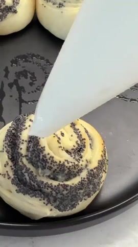 The video shows a close-up of a baking sheet with several round, swirl-shaped pastries dotted with poppy seeds. A white piping bag is held at an angle, dispensing a dollop of thick, white cream onto the center of one of the pastries. The cream is being carefully applied in a circular motion. The pastries appear to be golden brown and have a soft, doughy texture. The baking sheet is black and has a slightly wet, reflective surface, suggesting it may have been greased. The background is out of focus but appears to be a light-colored surface, possibly a countertop. The lighting is bright and focused on the pastry and piping bag.