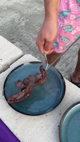 The video shows a partially sliced piece of cooked meat being lifted off a blue plate with a fork. A man wearing pink shorts with a crocodile pattern and black sandals can be seen in the video. He also picks up several skewers from a charcoal grill. The grill contains what appears to be meat of various shapes and sizes. The scene takes place on a concrete surface, presumably outdoors.