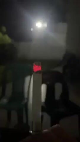 The video shows a lit cigarette held in someone's hand. The tip of the cigarette glows red, and smoke is rising from it. The background is dark with a light in the distance. There are two plastic chairs, one green and one blue, visible in the blurred background. The scene appears to be outdoors at night.