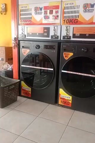 The video shows the interior of a laundromat. Two large black washing machines are visible in the foreground, each with a circular glass door. Stickers on the machines indicate they have a 10KG maximum load capacity. Above the washing machines, there are several dryers stacked vertically. A dark grey laundry basket sits to the left of the washing machines. In the background, a yellow wall is visible, and a portion of a street with motorcycles parked is seen through an open doorway. A person wearing a dark hijab and a light-colored jacket is sitting on the right side of the frame, looking at a tablet.