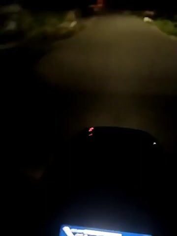 A first-person perspective video shows a motorcycle riding down a paved road at night. The motorcycle's headlight illuminates the road ahead, showing the gray pavement and grass on either side. Houses are visible in the distance, lit by dim streetlights. The wind noise is audible, indicating the motorcycle is in motion. The camera angle is from behind the handlebars, showing a small portion of the dashboard.