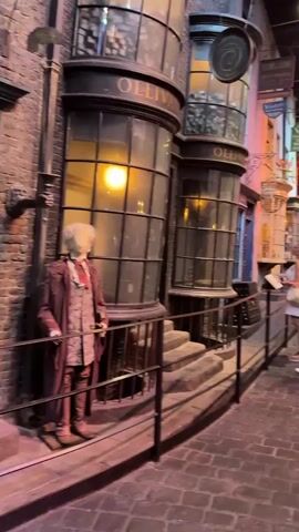 The video shows a busy, cobblestone street lined with tall, narrow, old-fashioned shops. The buildings have large, curved windows and ornate details. A mannequin dressed in a reddish-brown robe and white shirt stands outside one of the shops.

Several people are walking through the street, some looking at the shop windows. Two women are standing near the foreground, one holding a phone and the other a book. In the background, more people are visible, as well as other shops with brightly colored facades, including one with prominent orange and red accents. The scene is illuminated by artificial lighting, creating a warm and somewhat magical atmosphere.

Visible text includes "OLLIVAN..." on the shop in the foreground, and "BREWS & STUES" and "FRUIT & VEGETABLES" on signs above other shops. Another sign reads "SLUG & JIGGERS." The overall impression is of a lively, themed attraction, possibly a film set or a retail area designed to resemble a historic or fantastical street. The time of day is not discernible due to the indoor setting and artificial lighting. The weather is irrelevant as it appears to be an indoor environment. The location context is Three Rivers, United Kingdom.