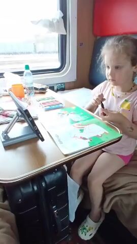 A young girl with blonde hair, wearing a pink t-shirt and pink shorts, sits on a train seat with a tray table in front of her. She is focused on coloring a book that is open on the tray table. The book has pictures of animals, and the girl is holding a marker or crayon in her right hand, appearing to be coloring one of the images. To her left, on the tray table, are a bottle of water, an orange cup, and a box of colored pencils. The train compartment has a window to the left, showing a blurry outdoor scene, and red and gray upholstery. A dark suitcase is visible below the tray table.