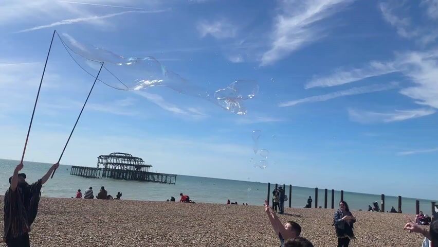 A man uses two poles to create giant soap bubbles on a pebble beach on a sunny day. The bubbles float in the air as people walk around and watch. Some people try to touch the bubbles. In the background is the ocean, a pier, and buildings. The sky is blue with streaks of clouds.