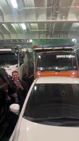 The video appears to be taken inside a ferry's vehicle deck. In the foreground, the hood and windshield of a white car are visible. To the left, a woman with blonde hair holds a phone and a water bottle. Behind the white car, an orange truck is parked, with people visible inside its cabin. Further back, a white van with a colored stripe and text on its side is parked. A man wearing a purple scarf stands near the van. The environment is dimly lit, with overhead lights and reflective surfaces. The camera moves from a frontal view to focus on the side of the white van, showing a person's arm extending from the driver's side window. The overall scene suggests the loading or unloading of vehicles onto a ferry.