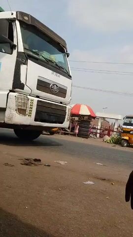 The video shows a white FAW truck parked on a paved street in what appears to be a bustling outdoor market. The front of the truck is prominently displayed, showing some wear and tear. In the background, an umbrella stall with tires stacked around it, a red and white fence, and a yellow three-wheeled vehicle can be seen. Debris and trash are scattered on the road in front of the truck. A horn honks, indicating the presence of traffic. A person's hand appears briefly in the lower right corner of the frame. The sky is overcast, suggesting a cloudy day.