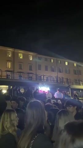 The video captures a lively outdoor night event in Turin, Italy, filled with a large crowd of young people. The setting appears to be an urban square or open area, framed by a multi-story building with illuminated windows, suggesting a classic European architectural style.

An elevated platform or balcony acts as the central stage for the event. Here, at least two individuals operate a DJ setup, surrounded by lights. A prominent neon sign on the platform glows brightly, displaying a stylized word resembling "VOGUE," with additional characters beneath it. Directly below this neon sign, white text clearly spells out "BAR."

The crowd, consisting predominantly of young adults, is densely packed, standing, and facing the elevated platform, indicating engagement with the music or performance. The dark sky confirms the nighttime setting, and there are no signs of adverse weather. The overall atmosphere is energetic and social, typical of an outdoor party or concert taking place under the night sky in a vibrant city.