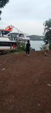A video shows a boat docked at a waterfront. The boat is white with orange accents and decorated with flags. People are visible on the boat, including some standing on deck and others seated inside. Several children are gathered on a wooden platform next to the boat. A few children are walking away from the camera towards the boat. The environment appears to be a natural setting with trees and hills in the background, and the water is calm. The ground in the foreground is covered with dirt and some debris. The sky is overcast.