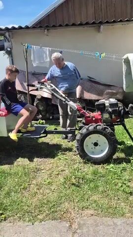 A young boy sits on a trailer attached to a tiller while an older man stands nearby, speaking to him. The boy is wearing a dark t-shirt with red and white accents, blue shorts, and yellow slides. He is looking towards the camera and smiling. The older man is wearing a blue t-shirt and khaki pants. He is holding his hand up to his face, as if shielding his eyes or speaking. The trailer is made of metal and has a red seat. The tiller is red and white with large wheels. The scene appears to be in a yard with green grass and a building in the background. There are clothes hanging on a clothesline in front of the building. The sky is blue with some clouds.