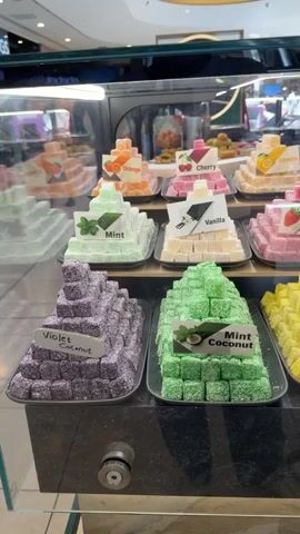 The video pans across a display case filled with pyramids of colorful Turkish delight. The display is divided into sections, each featuring a different flavor. From left to right, top to bottom, the visible flavors include Violet Coconut, Mint Coconut, Lemon Coconut, Orange, Cherry, Lemon, Rose, Vanilla, Strawberry, Banana, Pomegranate, Apple, and Cherry Coconut. The Turkish delight pieces are uniformly shaped and coated in either sugar, powdered sugar, or desiccated coconut. The display case is well-lit, and the background suggests a retail environment with other displays visible. There are no people visible in the video.
