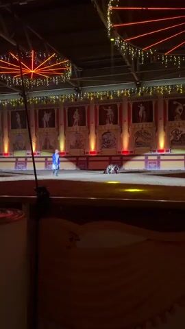 A man in a blue tunic and white cape, holding a sword, stands on a sand-covered stage. Another man, wearing armor and a red tunic, is lying on the ground in front of him. The man with the sword appears to be interacting with the fallen man. The background features a stage set with Roman-style columns and murals. String lights are draped across the ceiling. The scene is lit by spotlights.
