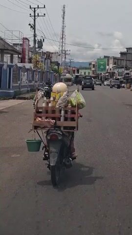 The video shows a person riding a motorcycle down a paved road in a town or city. The motorcycle has a large wooden crate attached to the back, which contains multiple bottles, a white round object, a bag of some sort and a green bucket hanging off the back. The rider is seen from the back and their legs are visible, clad in brown pants. Other vehicles can be seen on the road ahead, including cars and trucks. Buildings, streetlights, and power lines line the sides of the road. A tall cell phone tower is visible in the background. The sky is overcast. The general atmosphere is that of a bustling, developing urban area.