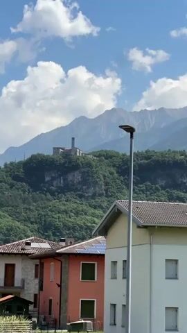 The video shows a town with a view of the mountains in the background. The camera pans across several houses with red-tiled roofs. A building that looks like a castle sits on top of a hill, surrounded by trees.  The sky is blue with white clouds.  The mountain range is silhouetted against the sky.
