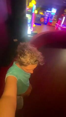 The video opens with a selfie-style shot of a person with curly, light brown hair wearing a teal t-shirt and grey shorts. They are in a roller skating rink with flashing arcade game lights in the background. The floor is a deep red. The person is moving forward, and the camera is looking down and to the side. As the video progresses, the person continues to skate, with other skaters visible in the background. The arcade games are a blur of neon colors. The camera movement is dynamic, capturing the motion of skating. The video appears to be shot from a first-person perspective while the person is actively participating in roller skating.